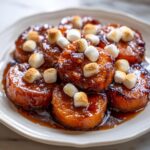 Close-up of glazed, round candied yams topped with toasted mini marshmallows on a white plate.