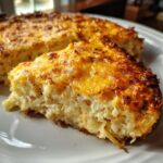 A close-up slice of baked cheesy potato casserole showing shredded potatoes and a golden, bubbly cheese crust.