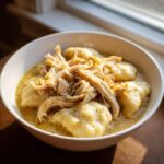 Close-up of a white bowl filled with creamy chicken and dumpling soup, topped with shredded chicken.