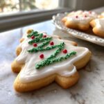 Close-up of a Christmas tree-shaped sugar cookie decorated with white royal icing and green sprinkles.