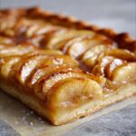 Close-up, mouthwatering shot of a slice of glistening apple tart showing caramelized apple slices on a shortbread crust.