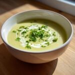 A bowl of creamy, pale green celery soup garnished with a swirl of cream and fresh chopped parsley.