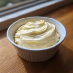 A small white ramekin filled with thick, creamy homemade mayonnaise, sitting on a wooden surface.