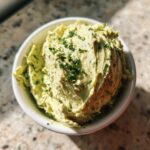 Overhead close-up of a creamy, light green olive dip topped with fresh chopped herbs in a white bowl.
