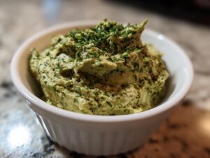A close-up shot of a creamy, textured green olive dip, garnished with fresh herbs, served in a small white ramekin.