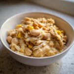 A close-up of a white bowl filled with creamy white chicken chili, featuring shredded chicken, white beans, and yellow corn kernels.