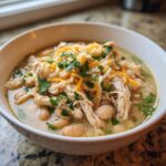 A close-up of a bowl of creamy crockpot white chicken chili topped with shredded cheese and fresh cilantro.