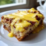 A close-up of a slice of eggs benedict casserole topped generously with rich, bright yellow hollandaise sauce.