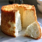 A tall, freshly baked angel food cake with a slice cut out, showing its airy white texture.
