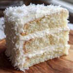 Close-up of a moist, fluffy slice of three-layer coconut cake covered in white frosting and shredded coconut.