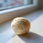 A smooth, pale yellow ball of freshly prepared marzipan recipe dough resting on a white surface near a window.