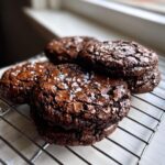 A stack of rich, fudgy brownie cookies topped with flaky sea salt cooling on a wire rack.