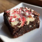 Close-up of a fudgy candy cane brownie square topped with white frosting and crushed peppermint pieces.
