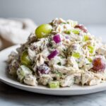 A mound of creamy greek yogurt chicken salad mixed with red onion, celery, and red and green grapes on a white plate.