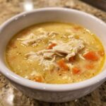 A close-up of a white bowl filled with rich, creamy cream of chicken soup featuring shredded chicken and diced carrots.