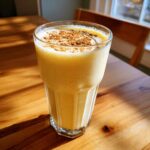 A tall glass of creamy, yellow homemade eggnog topped with a dusting of cinnamon, sitting on a sunlit wooden table.