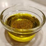 Close-up of a homemade salad dressing, featuring yellow oil and dark spices, in a small clear glass bowl.