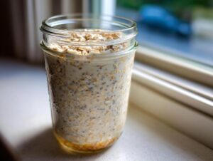A glass jar filled with creamy overnight oats mixed with chia seeds, sitting on a windowsill.