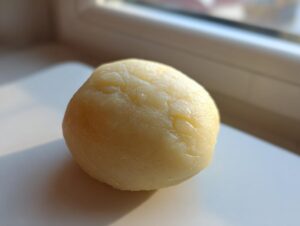 A smooth, pale yellow ball of homemade marzipan recipe dough resting on a white surface near a window.