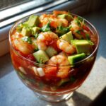 Close-up of a glass filled with vibrant mexican shrimp cocktail, topped with avocado and cilantro.