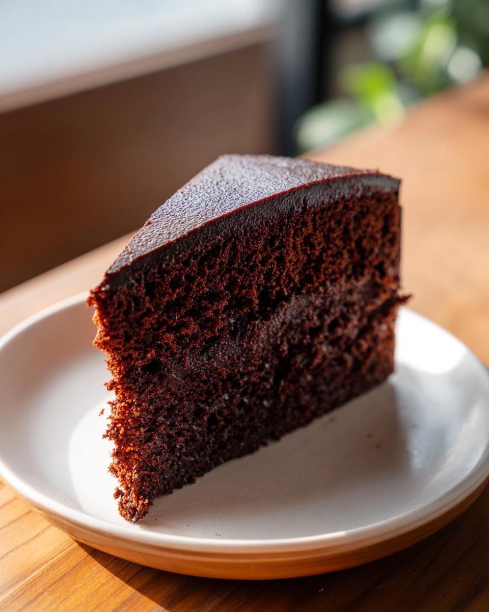 A rich, dark slice of moist chocolate cake with a smooth ganache layer on top, served on a white plate.