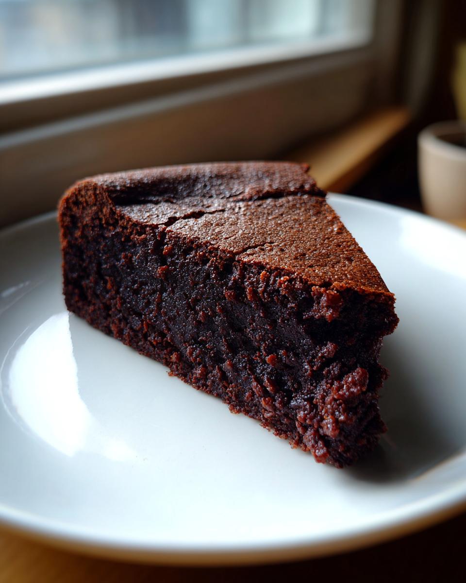 A close-up of a rich, dark slice of moist chocolate cake served on a white plate near a window.