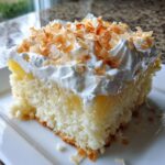 A close-up of a moist slice of coconut poke cake topped with whipped cream and toasted coconut flakes.