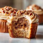 A pecan pie cupcakes with a bite taken out, revealing a gooey pecan filling and topped with brown sugar frosting and chopped pecans.