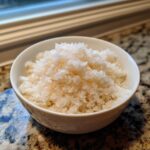 Close-up of perfectly seasoned sushi rice glistening in a small white bowl on a granite countertop.