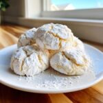 A stack of soft, crinkle-topped cool whip cookies heavily dusted with powdered sugar on a white plate.
