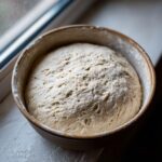 Close-up of perfectly proofed pizza dough recipe rising in a brown bowl near a window.