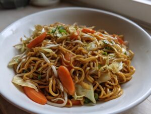 A close-up of a white bowl filled with savory fried noodles mixed with sliced carrots, cabbage, and bean sprouts.