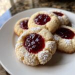 A plate holding several delicious raspberry thumbprint cookies dusted lightly with powdered sugar.