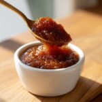 A spoonful of thick, dark brown pear butter is being lifted from a small white ramekin.