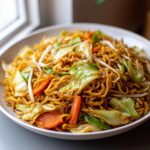 A close-up shot of a bowl filled with glossy, brown fried noodles mixed with crisp cabbage, carrots, and bean sprouts.