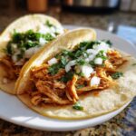 Close-up of two soft corn tortillas filled with shredded chicken tacos, topped with diced white onion and fresh cilantro.
