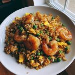 A close-up of a white bowl filled with delicious shrimp fried rice recipe, featuring large shrimp, peas, and scrambled egg.