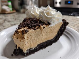 A close-up of a slice of creamy peanut butter pie with a dark chocolate crust, topped with whipped cream and cookie crumbs.