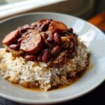 A close-up serving of slow cooker red beans and rice topped with slices of smoked sausage on a white plate.