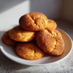 A stack of freshly baked, soft snickerdoodle cookies generously coated in cinnamon sugar, resting on a white plate.