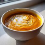 Close-up of a bowl of vibrant orange squash soup topped with a swirl of cream and pepper.