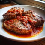 Three seared steaks covered in rich, red tomato sauce and herbs, known as Steak Pizzaiola, served on a white plate.