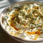 Close-up of creamy whipped goat cheese topped with fresh herbs, olive oil, and orange zest.