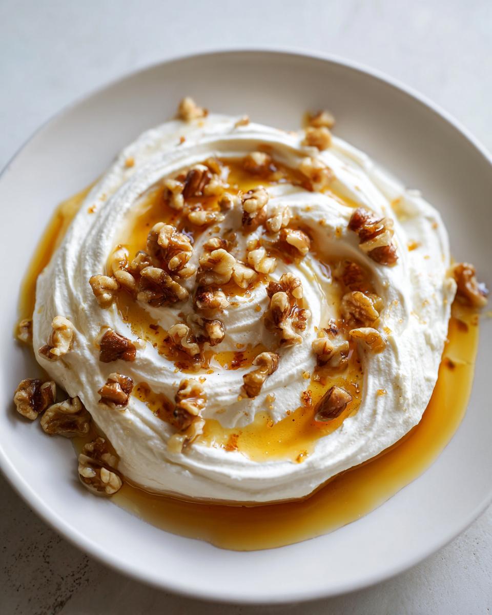 Close-up of a creamy whipped ricotta dip swirled on a white plate, drizzled with honey and topped with chopped walnuts.