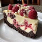 A perfect slice of white chocolate raspberry cheesecake with a dark crust, topped with fresh raspberries and white chocolate shavings.