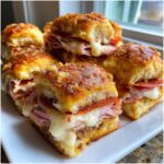 Close-up of several baked grinder sliders stacked on a white plate, featuring melted cheese, ham, and pepperoni.