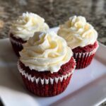 Three perfectly frosted red velvet cupcakes sitting on a white plate, featuring vibrant red cake and thick swirls of white cream cheese frosting.