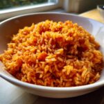 Close-up of a white bowl filled with vibrant, orange-red Jollof rice, ready to eat.