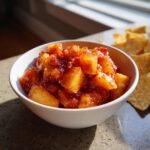 Close-up of a white bowl filled with chunky, glistening peach salsa next to tortilla chips.