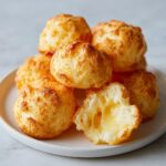 A stack of golden-brown Brazilian cheese bread puffs, with one broken open showing the gooey, melted cheese interior.